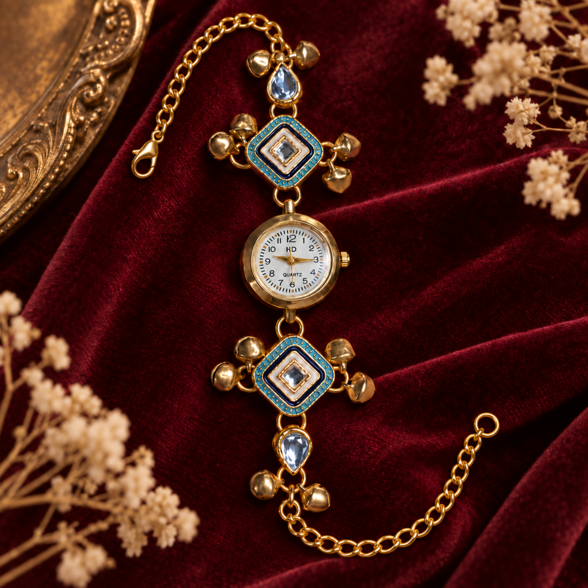 A gold-plated watch with turquoise enamel accents and small blue teardrop crystals, displayed on a deep red velvet fabric with dried white flowers.
