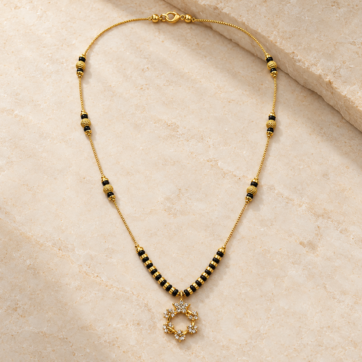 A gold chain necklace with black and gold beads, featuring a central gold ring pendant embellished with small clear stones, displayed on a light beige stone surface.