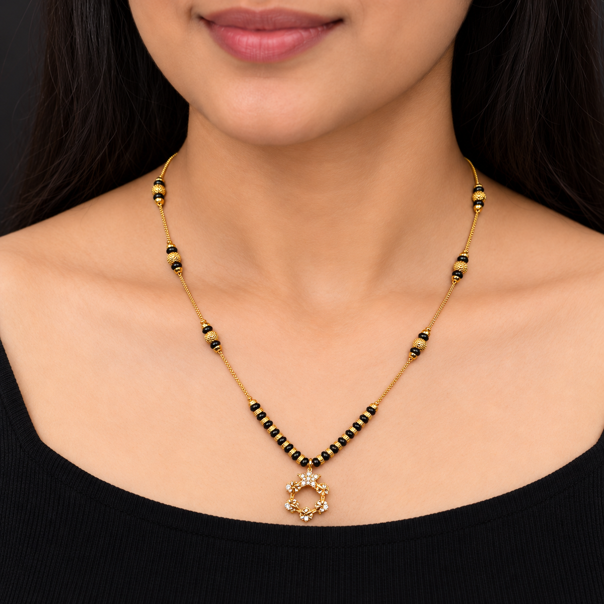 A close-up of a woman wearing a gold necklace with black bead accents and a small central pendant, shown against her neck and upper chest.