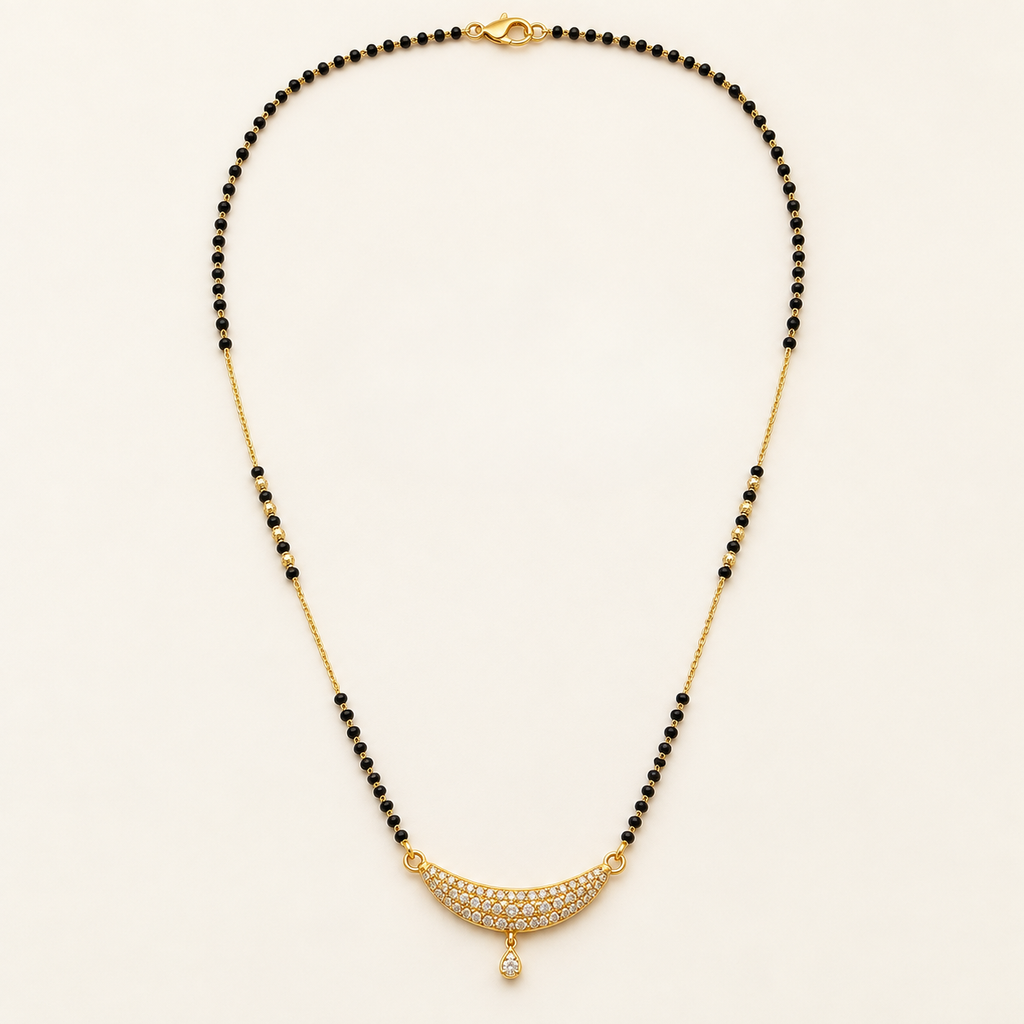 A gold and black beaded necklace with a crescent-shaped pavé pendant set with clear stones and a teardrop drop, displayed on a light cream background.