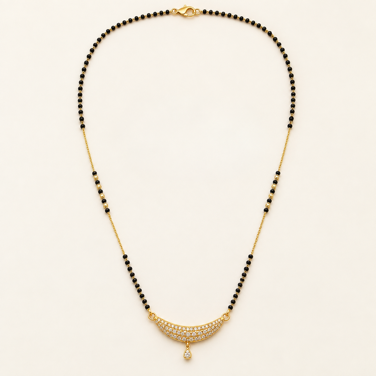 A gold and black beaded necklace with a crescent-shaped pavé pendant set with clear stones and a teardrop drop, displayed on a light cream background.