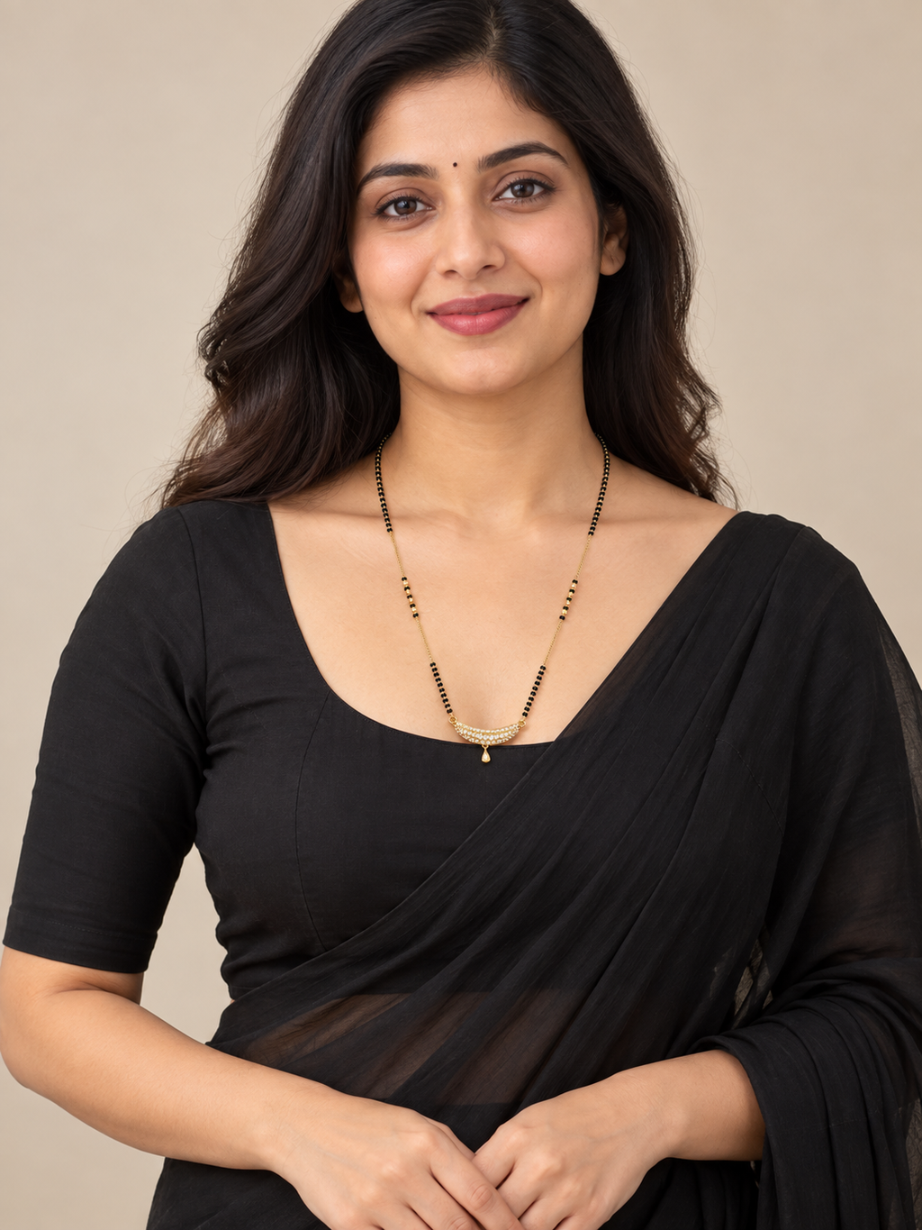 A woman with long dark hair wearing a black draped blouse and a delicate gold necklace, posing against a solid beige background.