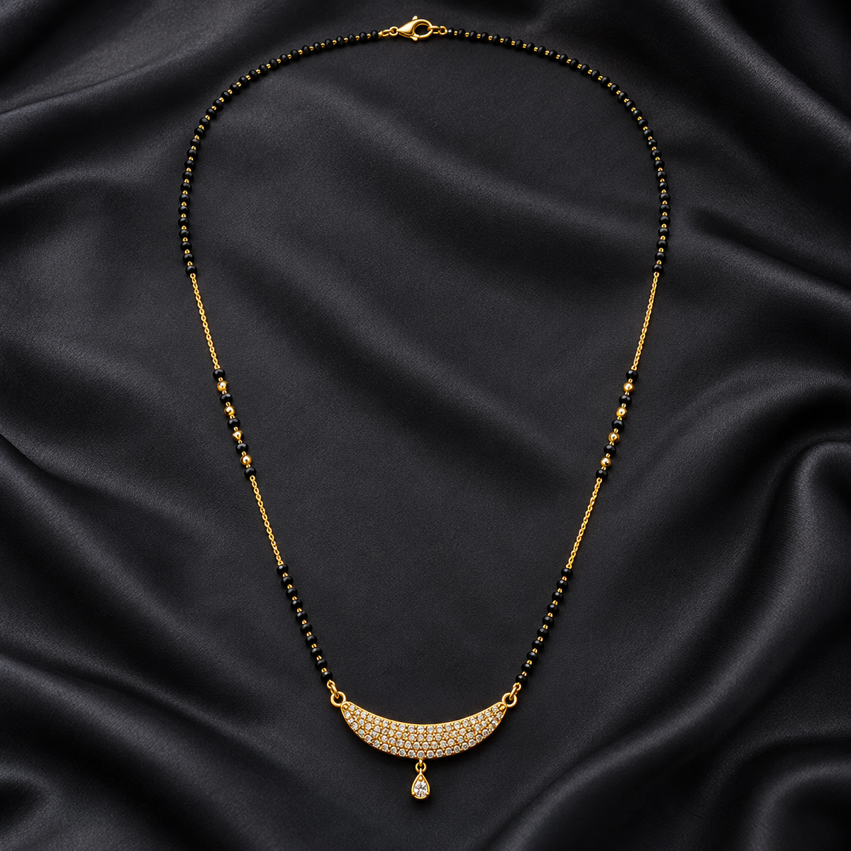 A gold-tone necklace with a crescent-shaped pavé pendant and a teardrop drop, laid out on a draped black satin fabric background.