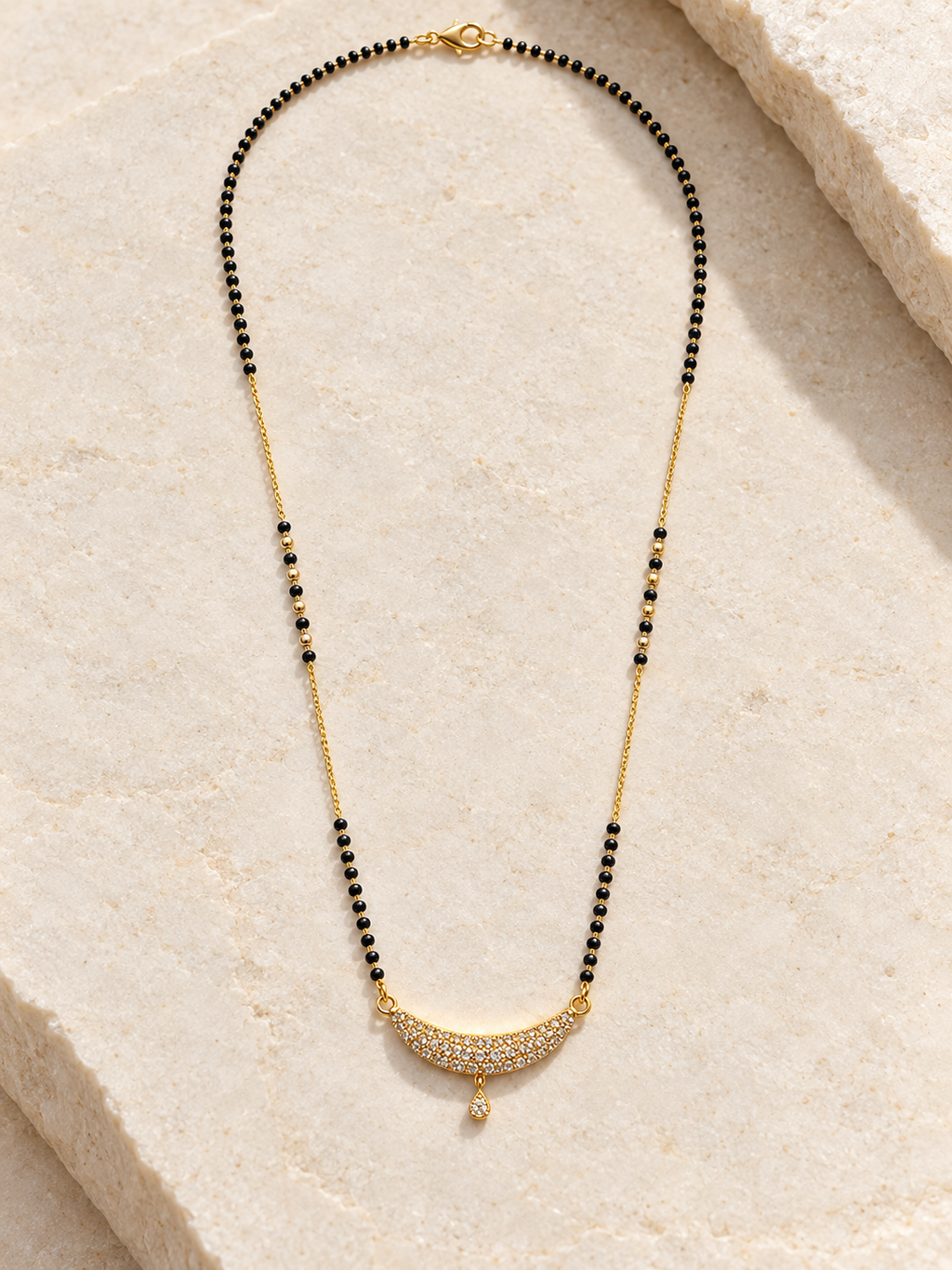 A gold and black beaded necklace with a crescent-shaped pavé pendant, displayed on a light beige stone surface.