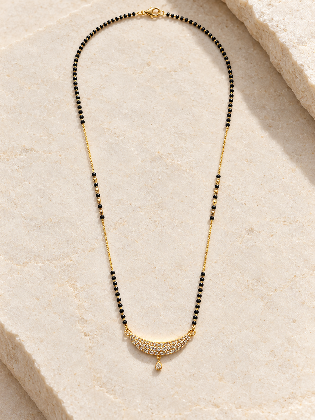 A gold and black beaded necklace with a crescent-shaped pavé pendant, displayed on a light beige stone surface.
