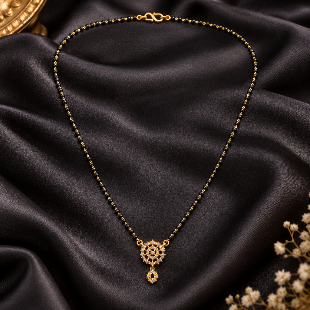 A gold-toned beaded necklace with a detailed circular pendant and teardrop charm, displayed on draped black satin fabric with small dried flowers in the corner.