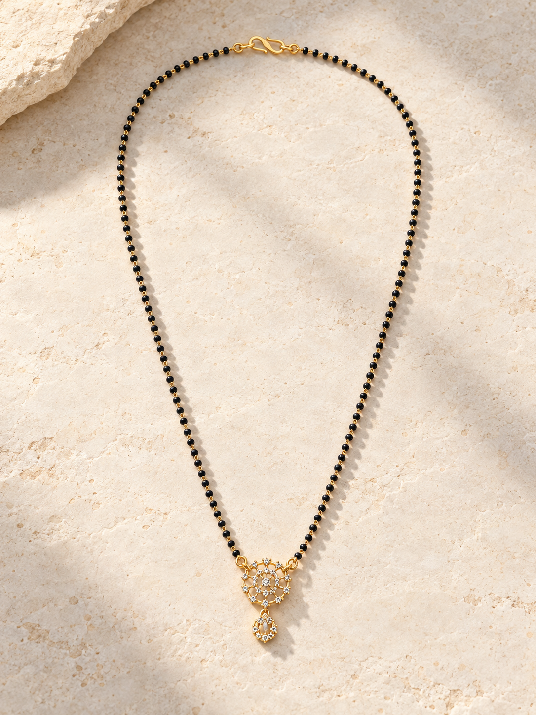 A beaded necklace with small black beads and a gold-toned pendant featuring clear stones, laid flat on a light beige textured stone surface.