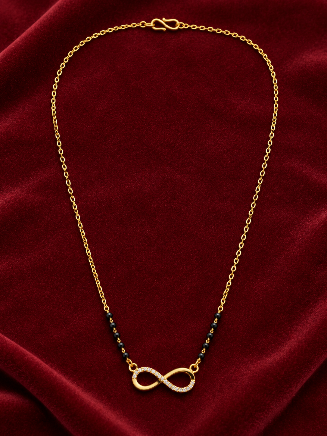 A gold chain necklace with a central infinity symbol pendant, accented by small dark blue beads on either side, displayed on a textured dark red velvet background.