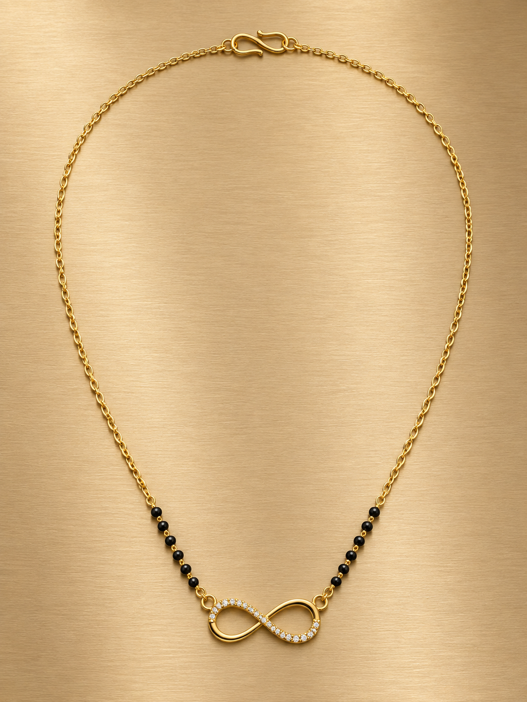 A gold chain necklace with a central infinity symbol pendant, accented with small black beads on either side, displayed on a brushed gold background.