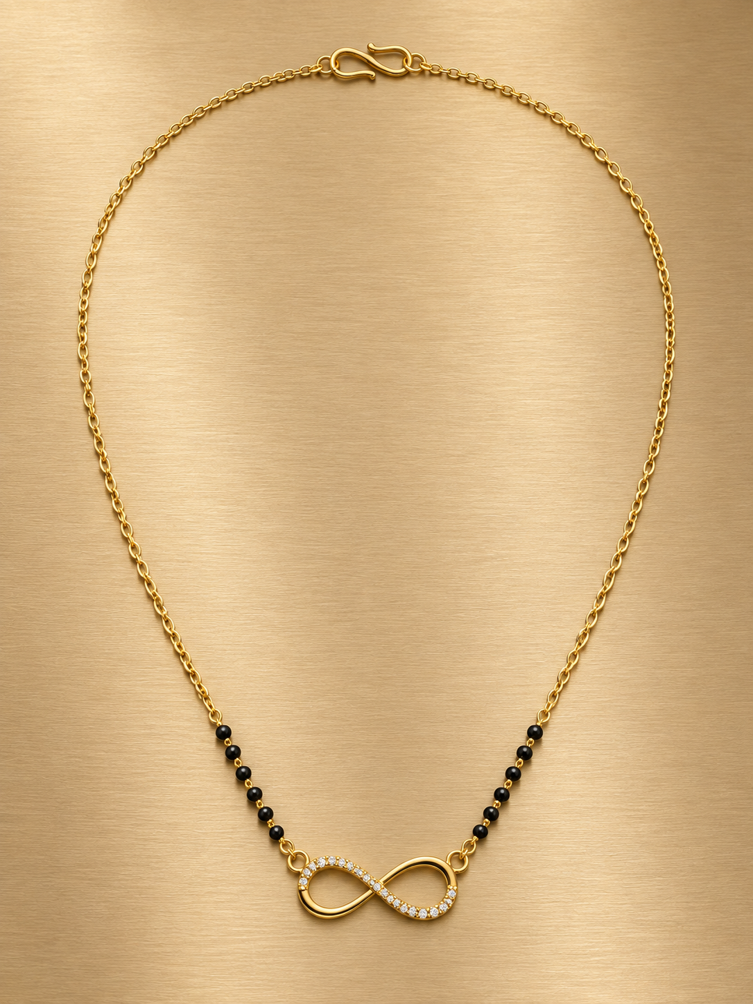 A gold chain necklace with a central infinity symbol pendant, accented with small black beads on either side, displayed on a brushed gold background.