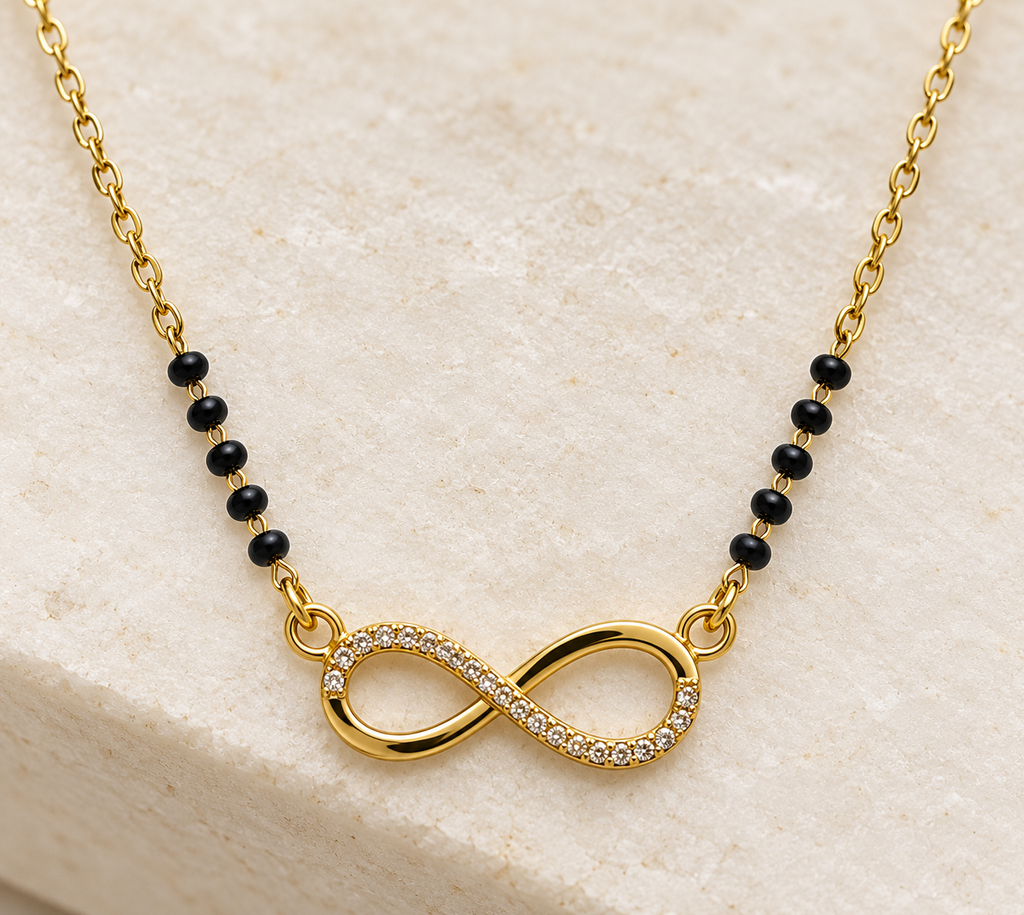 A gold chain necklace featuring a central infinity symbol paved with small clear stones, connected by black beads on either side, displayed on a light beige stone surface.