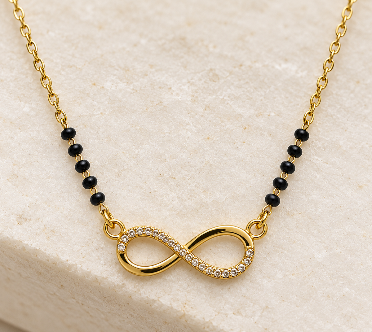 A gold chain necklace featuring a central infinity symbol paved with small clear stones, connected by black beads on either side, displayed on a light beige stone surface.