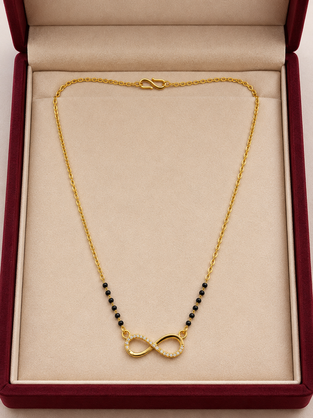 A gold chain necklace with a central infinity symbol pendant, accented with small black beads on either side, displayed inside a red velvet jewelry box with a beige lining.