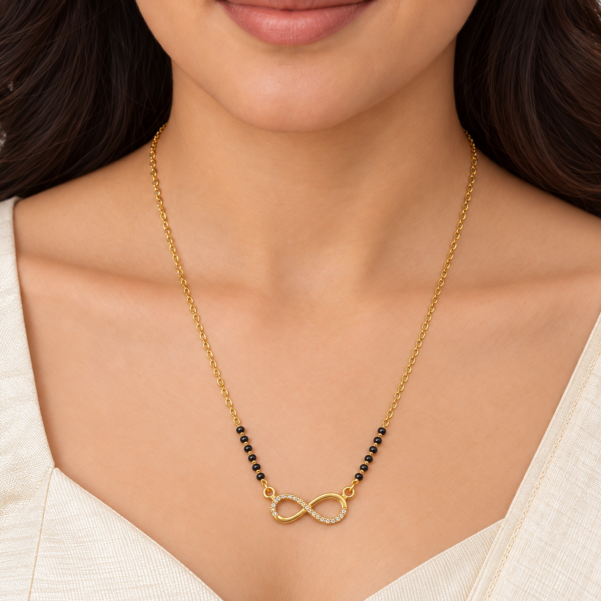 A close-up of a woman wearing a delicate gold chain necklace with a small infinity symbol pendant accented by tiny clear stones and black beads on either side, paired with a cream-colored top.
