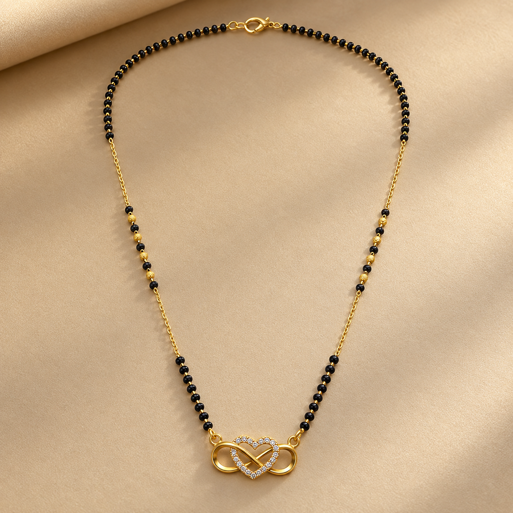 A gold chain necklace with small black beads and a gold infinity symbol pendant encrusted with clear stones, displayed on a beige fabric background.