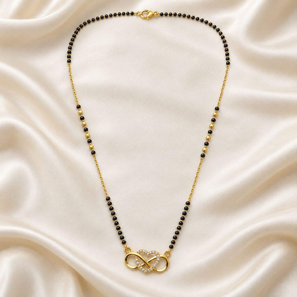 A gold chain necklace with small black beads and a gold infinity pendant accented with clear crystals, displayed on a draped cream satin fabric background.