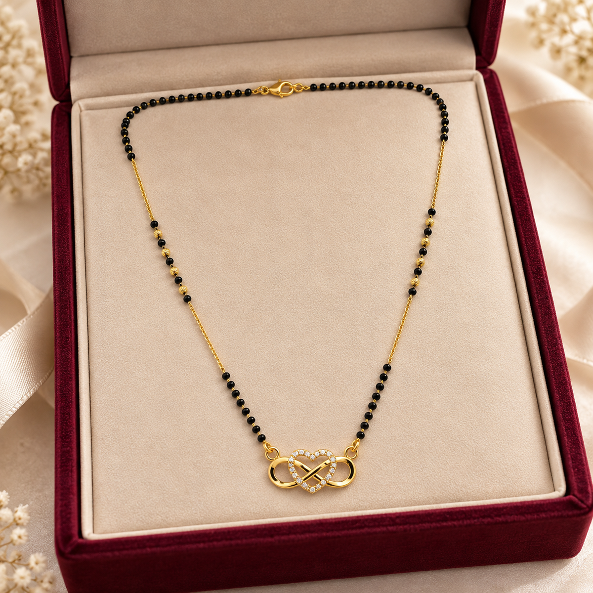 A gold necklace with small black beads and a central infinity symbol pendant accented with clear crystals, displayed inside an open burgundy velvet jewelry box with a beige insert.