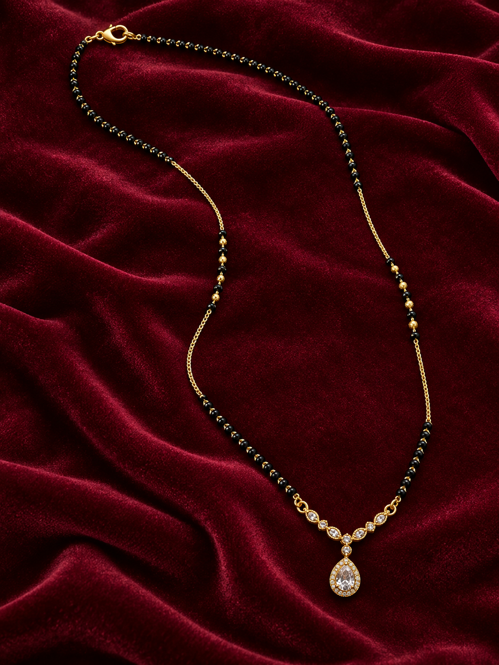 A gold chain necklace with small dark green beads and a teardrop pendant set with clear stones, displayed on a draped deep red velvet fabric.