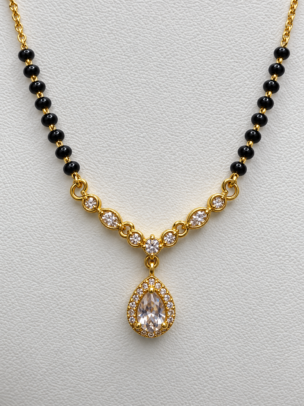 Gold and black beaded necklace with a teardrop-shaped clear crystal pendant, displayed on a textured light gray background.