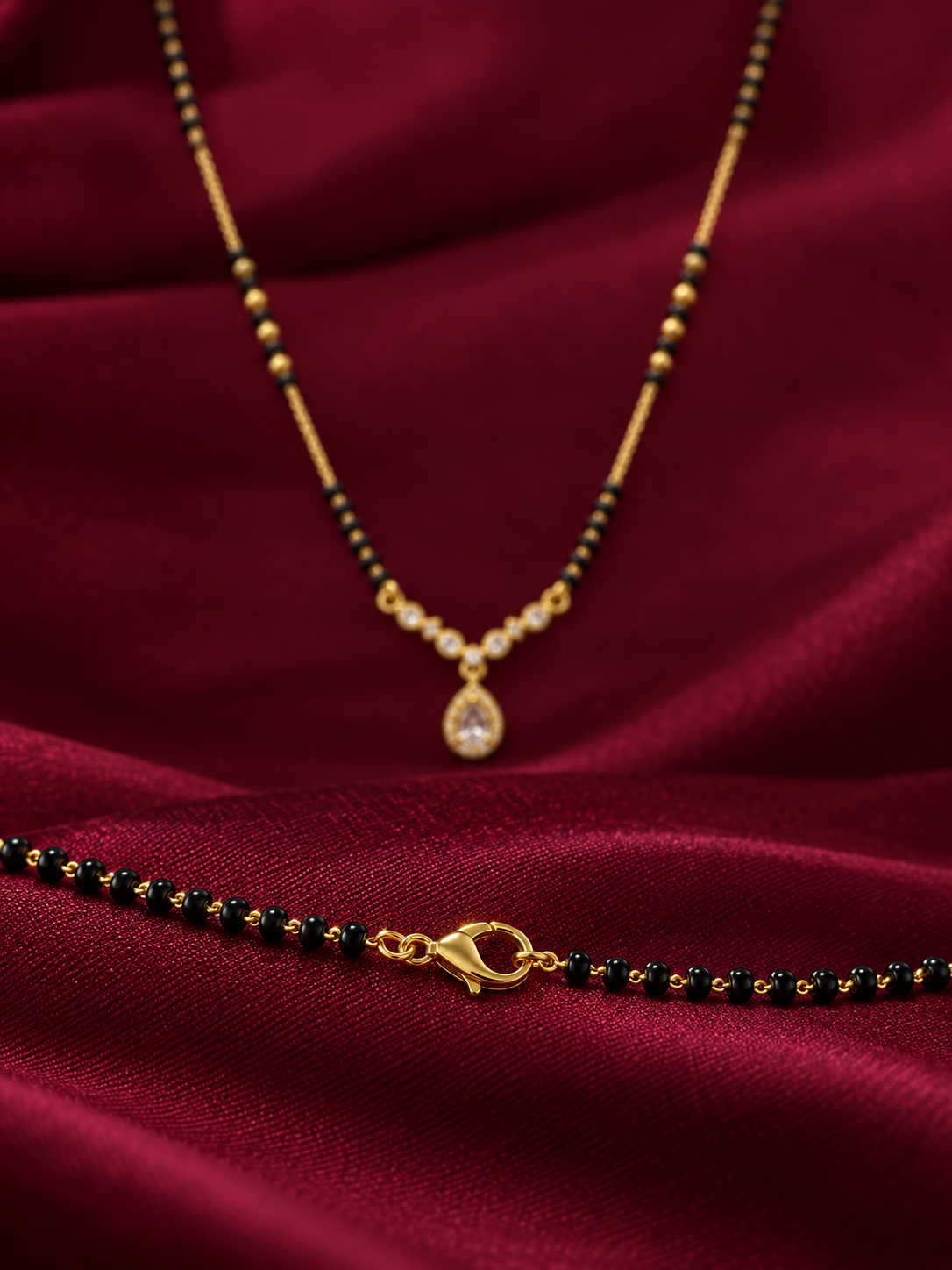 Gold beaded necklace with a teardrop-shaped clear stone pendant, displayed on draped deep red satin fabric alongside a matching beaded bracelet.