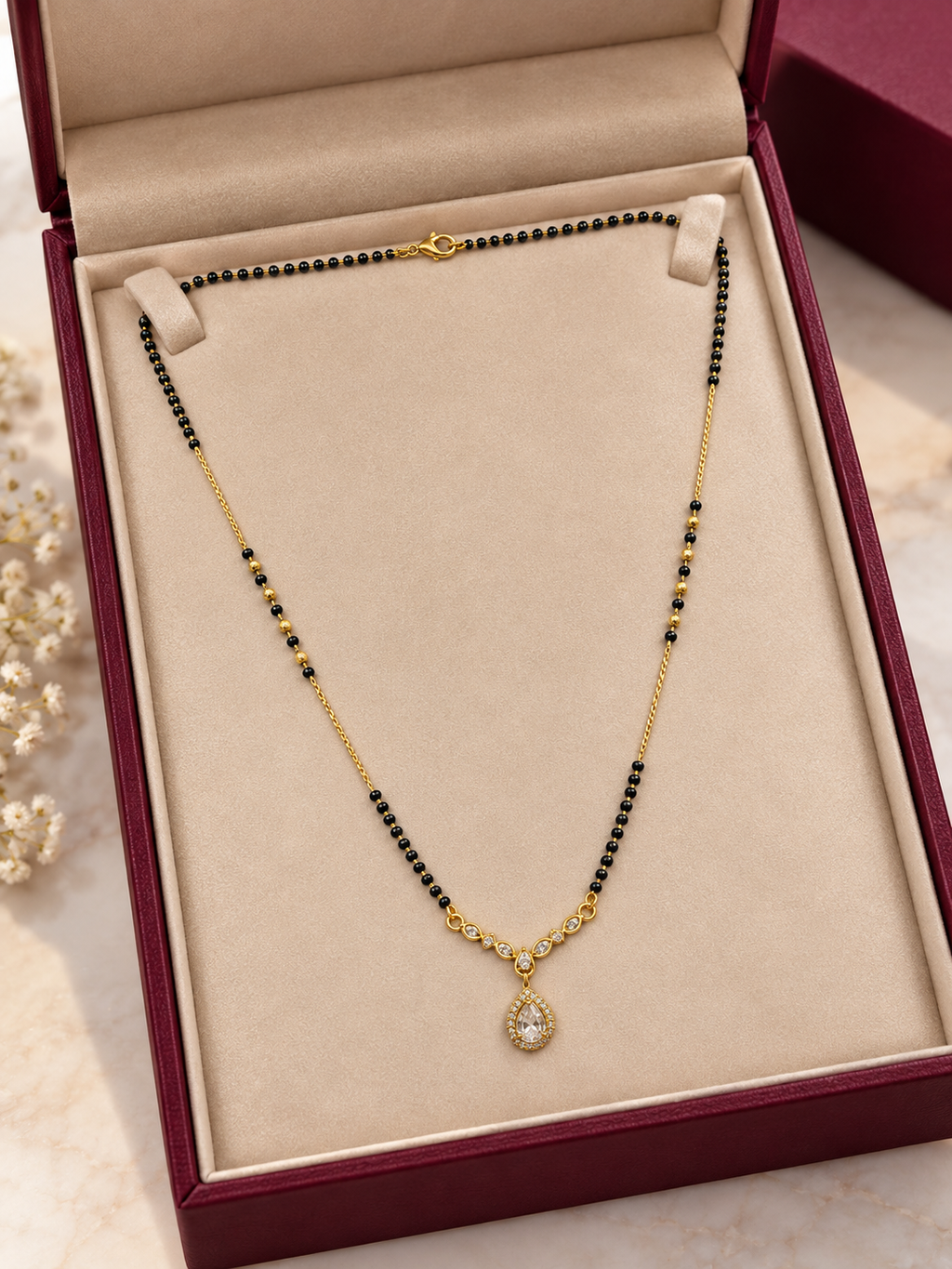 A gold and black beaded necklace with a teardrop-shaped clear stone pendant, displayed inside an open burgundy jewelry box with a beige velvet lining.