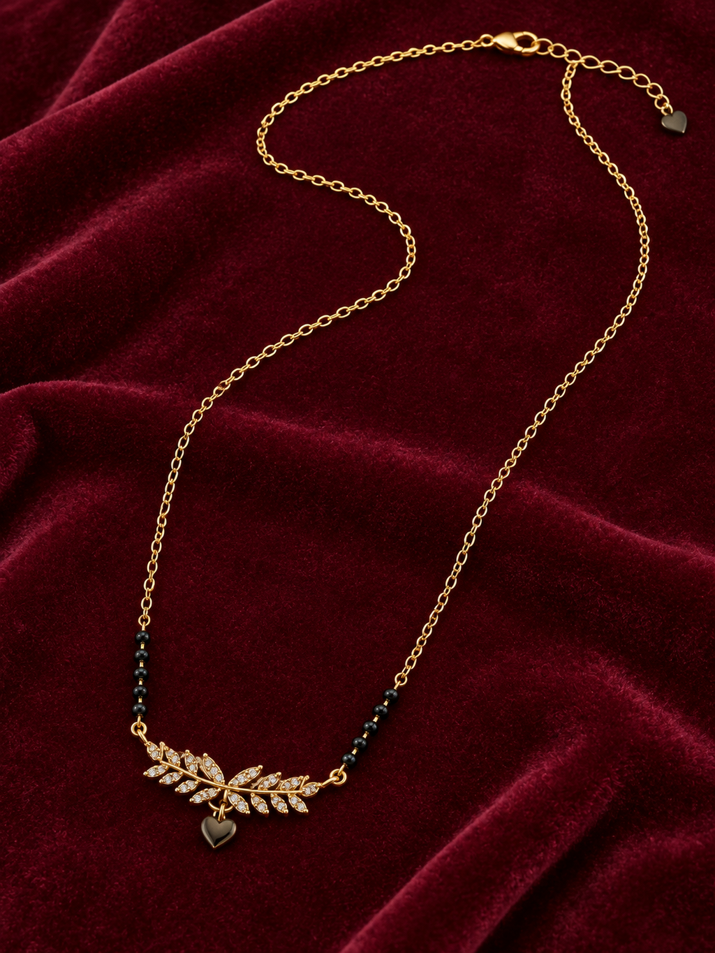 A gold chain necklace with a leaf-shaped crystal pendant and small dark heart charms, displayed on a deep red velvet fabric background.