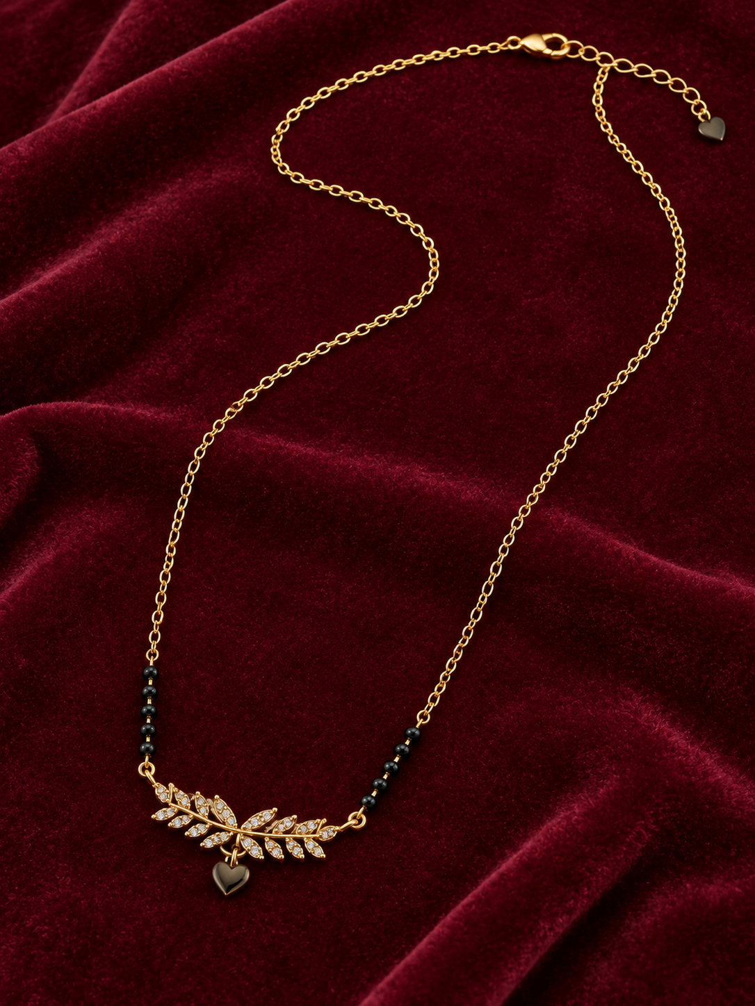 A gold chain necklace with a leaf-shaped crystal pendant and small dark heart charms, displayed on a deep red velvet fabric background.