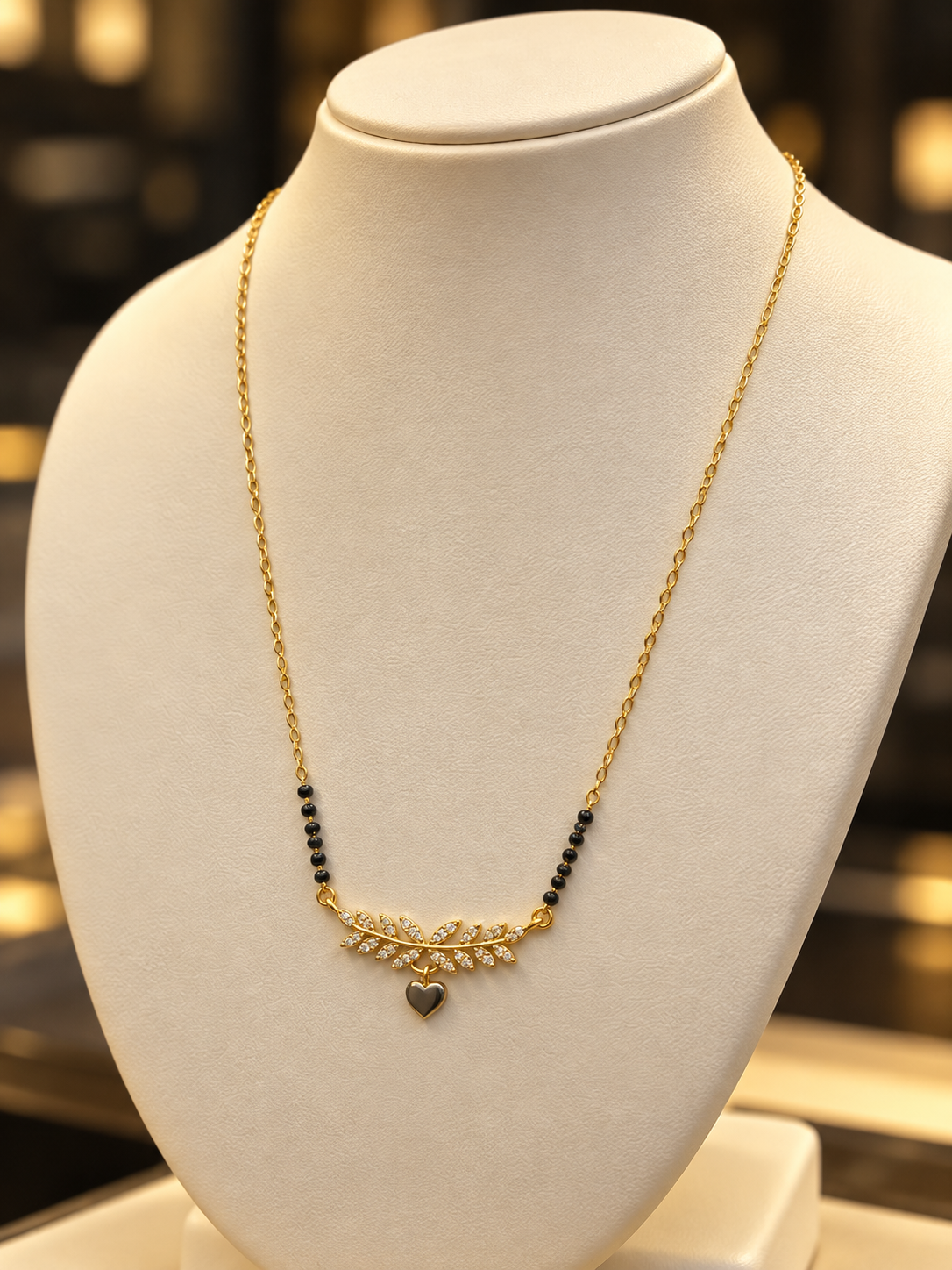 A delicate gold chain necklace displayed on a white jewelry bust. The necklace features a central gold leaf or branch motif adorned with small clear stones, flanked by small black beads on each side, and a small heart-shaped pendant hanging below.