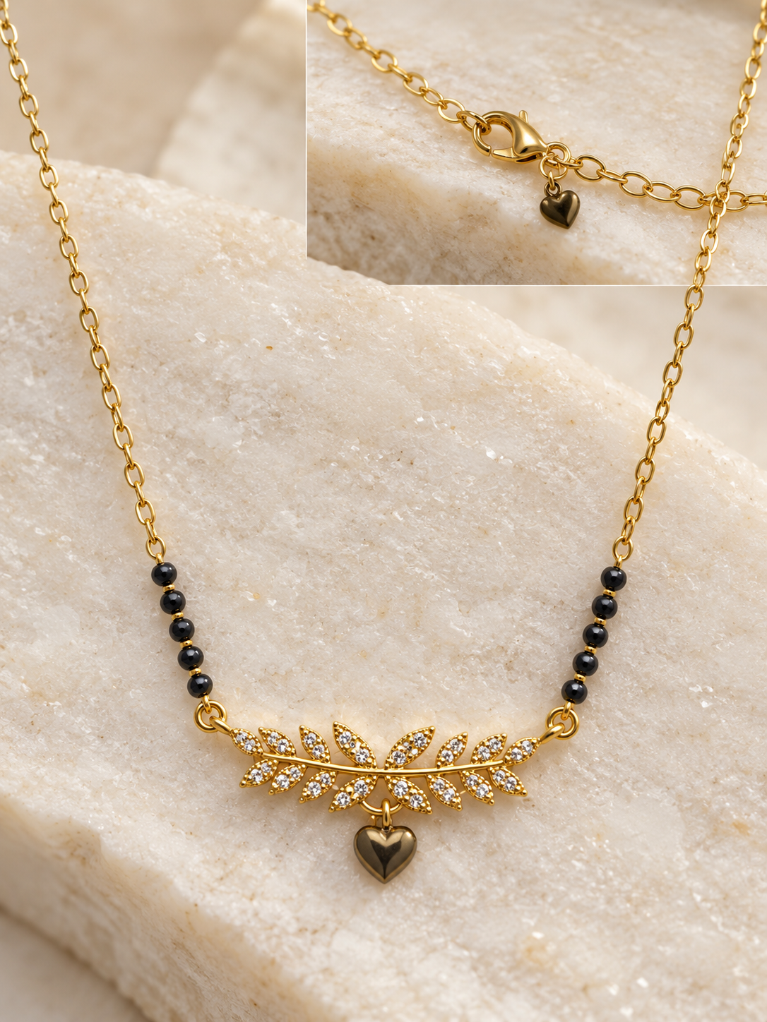 Gold chain necklace with a central leaf-shaped charm set with small clear stones and a dangling heart pendant, accented by black beads on each side, displayed on a light stone surface with an inset close-up of the clasp.