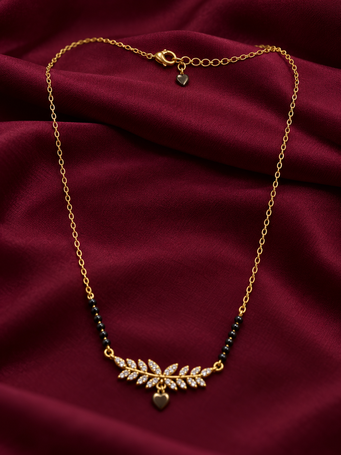 A gold leaf pendant necklace with small clear stones and a dangling heart charm, displayed on draped burgundy satin fabric.
