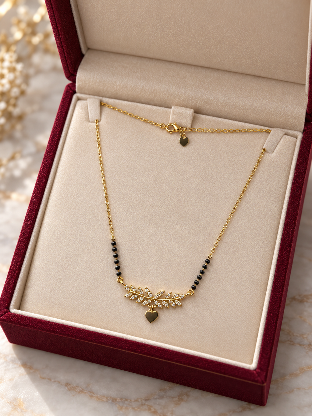 A delicate gold necklace with a leaf-like pendant, small black beads, and heart charms, displayed inside an open burgundy jewelry box with a cream insert.