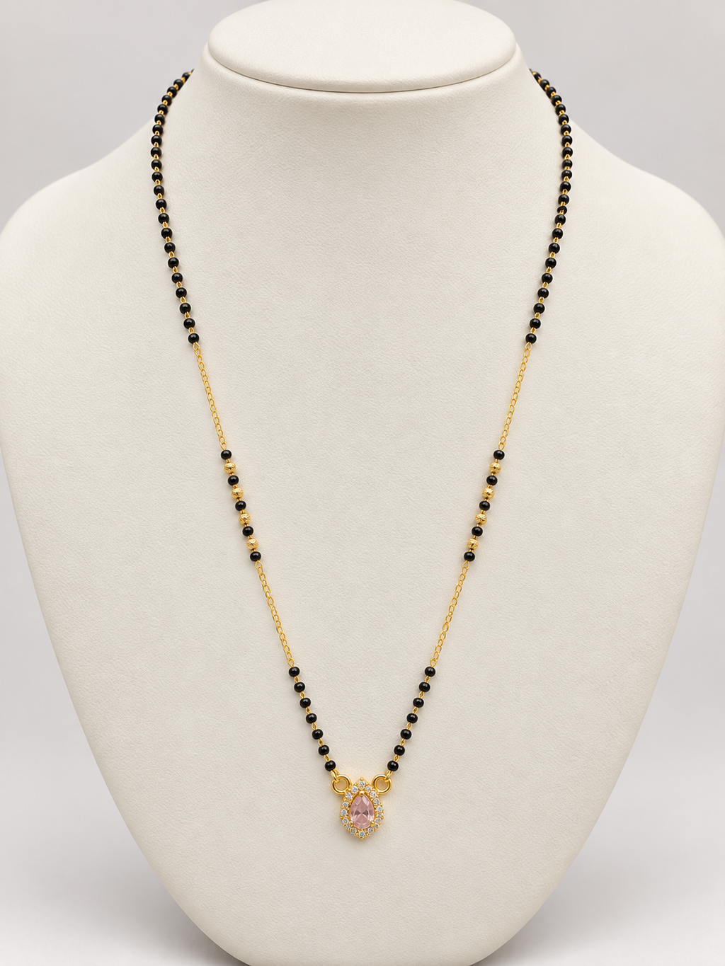 A gold chain necklace with small black beads and a central gold pendant featuring a pink gemstone, displayed on a white jewelry bust.