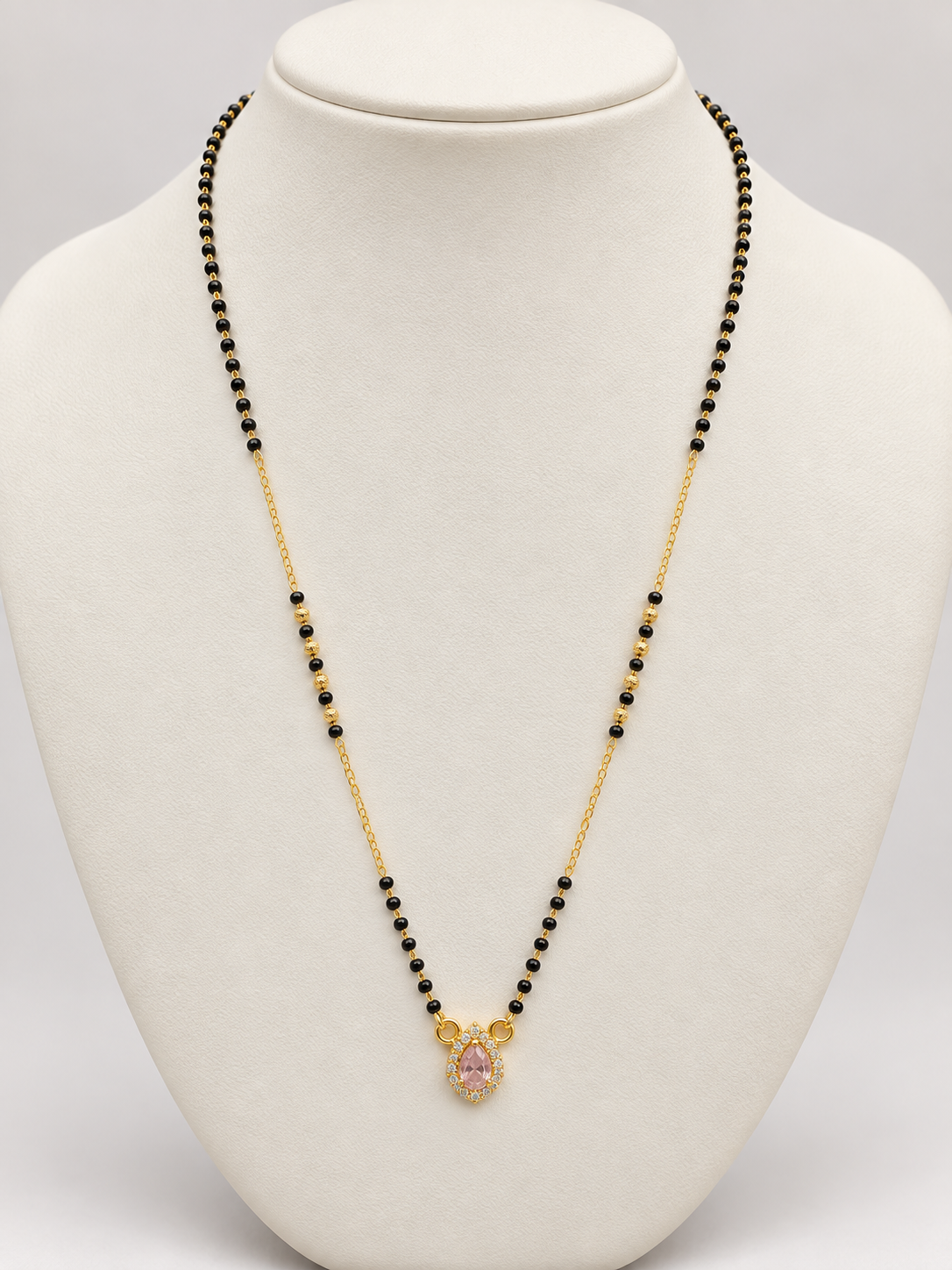 A gold chain necklace with small black beads and a central gold pendant featuring a pink gemstone, displayed on a white jewelry bust.