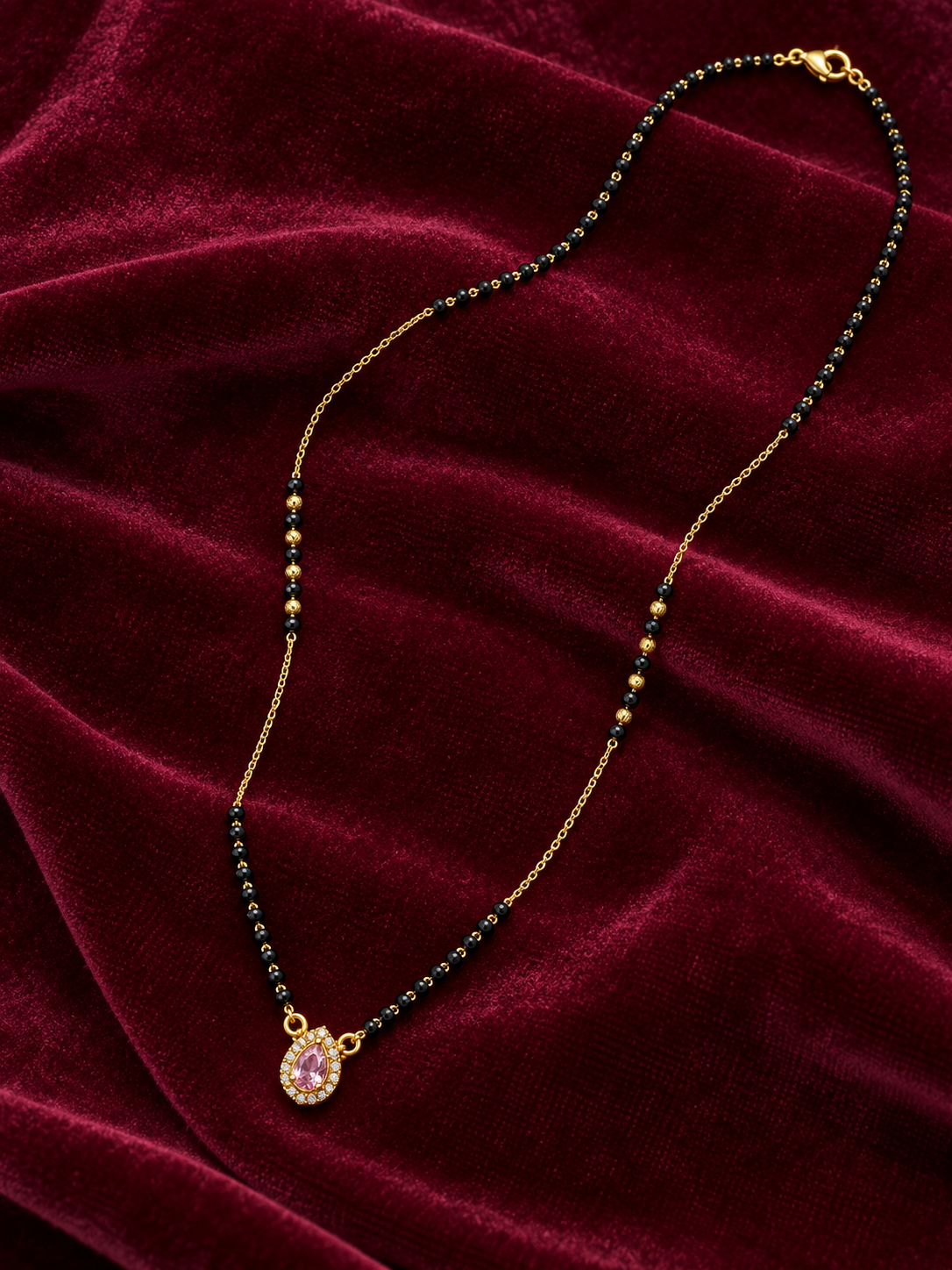 A gold chain necklace with small dark green and black beads and a teardrop-shaped pendant featuring a pink stone, displayed on a deep red velvet background.