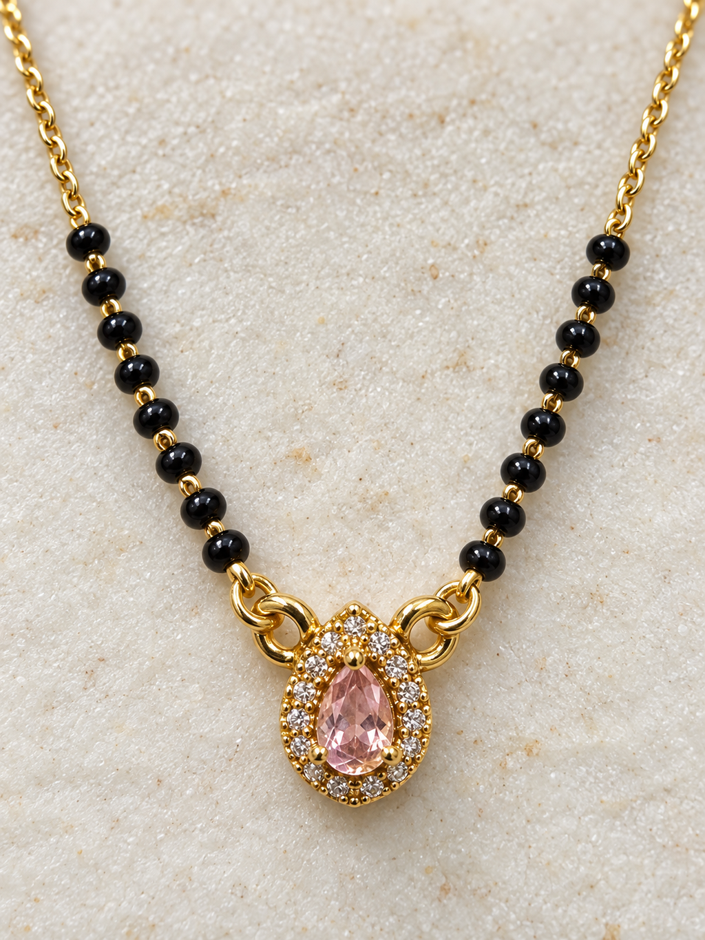 Gold chain necklace with small black beads and a teardrop-shaped pink gemstone pendant surrounded by clear stones, displayed on a textured light beige surface.