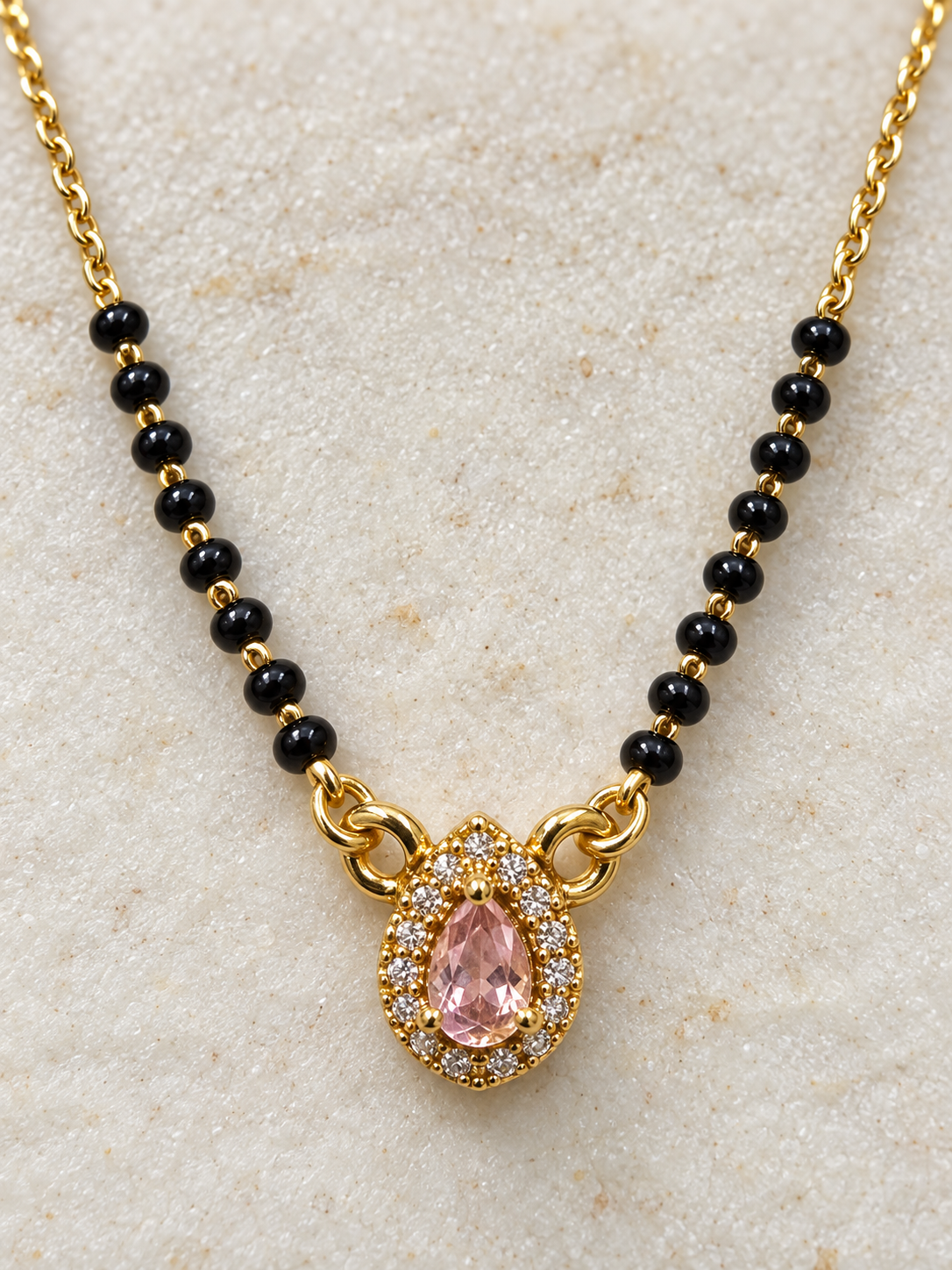 Gold chain necklace with small black beads and a teardrop-shaped pink gemstone pendant surrounded by clear stones, displayed on a textured light beige surface.