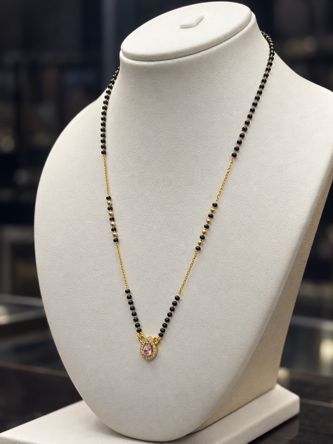 A gold and black beaded necklace with a small pink heart pendant, displayed on a white jewelry bust.