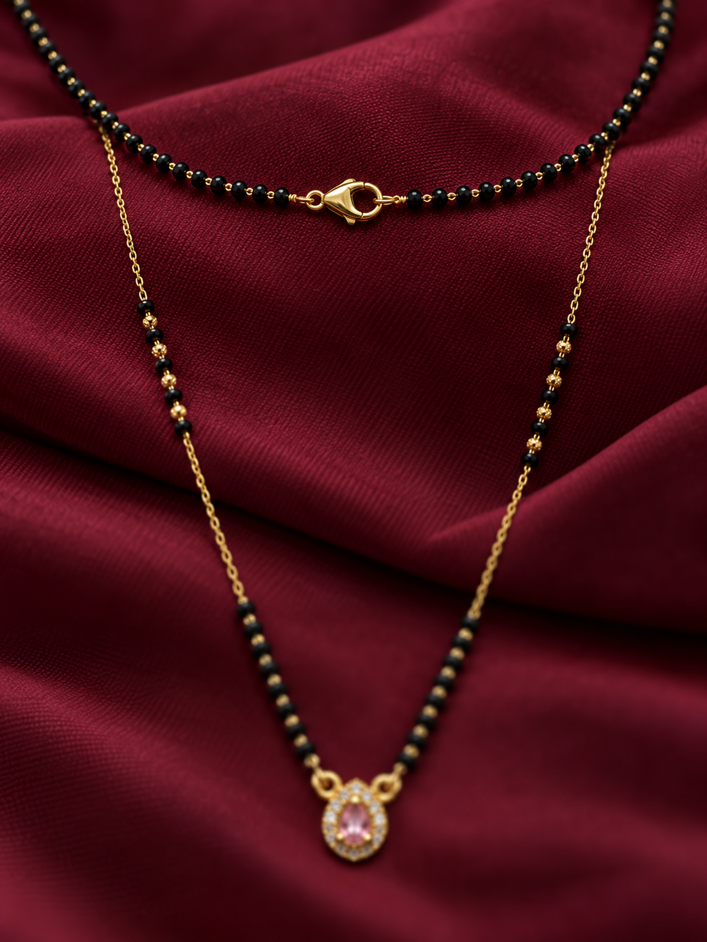 A gold chain necklace with a black beaded section and a teardrop-shaped pink gemstone pendant, displayed on draped dark red satin fabric.