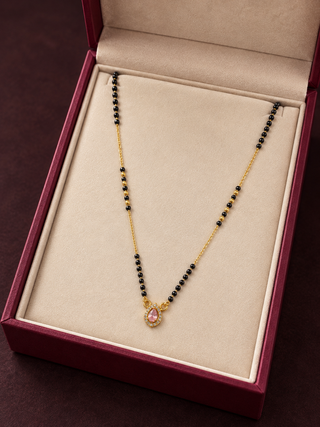 A gold and black beaded necklace with a pink teardrop pendant, displayed inside an open burgundy jewelry box with a beige lining.